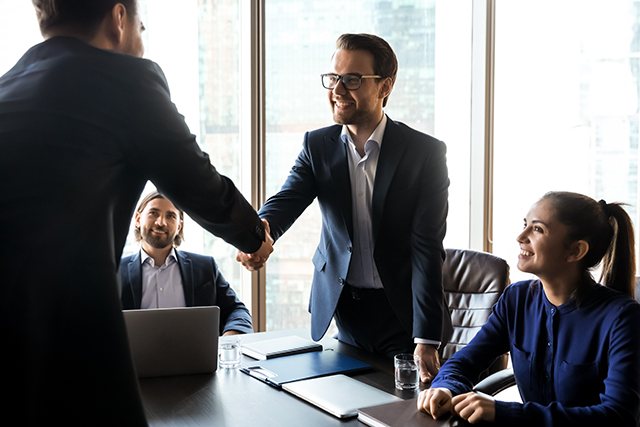 Happy successful businessman in suit shaking hand of applicant at interview. HR manager making good first impression. Employer congratulates attractive happy male candidate getting hired.